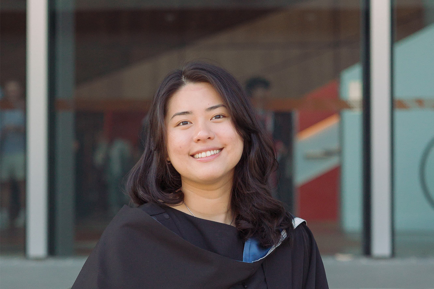 Angelina wearing a graduation gown, smiling for a photo in front of a large set of glass doors