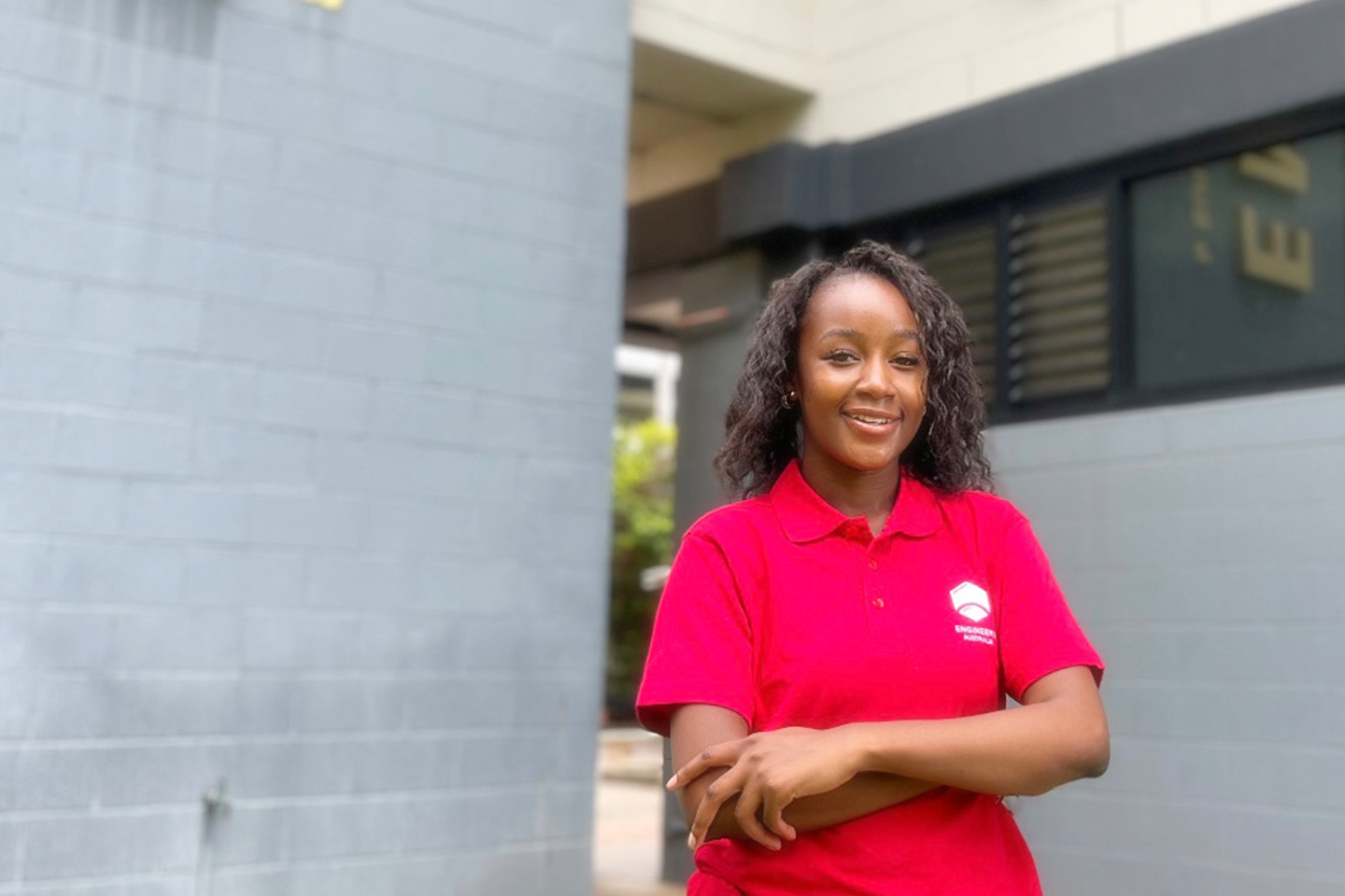 Elsie smiling at the camera as she leans against a grey concrete wall, shes wearing a bright red Engineers Australia polo shirt